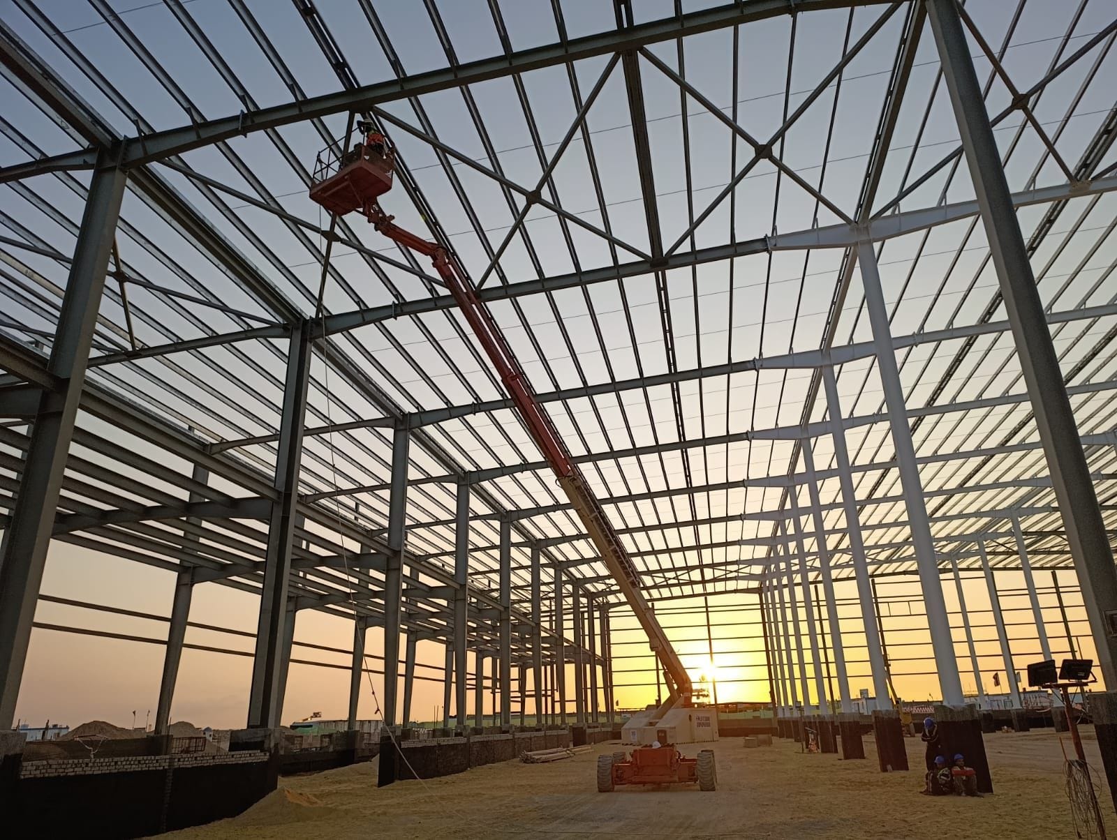 Steel erection crew on a boom lift installing roof steel inside the medical warehouse frame at sunset